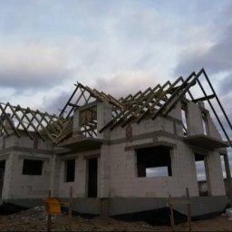 Michał Olechnowicz - Surowy stan domu jednorodzinnego z widoczną konstrukcją drewnianą dachu, w tle pochmurne niebo.