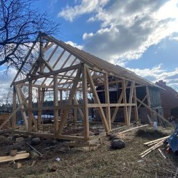 Michał Olechnowicz - Wznoszenie drewnianej konstrukcji szkieletowej budynku z widoczną więźbą dachową na tle błękitnego nieba z chmurami, w otoczeniu drzew i materiałów budowlanych.
