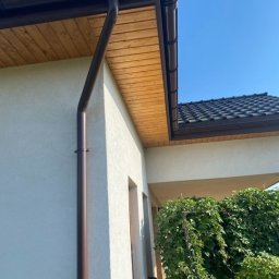 Michał Olechnowicz - Dom jednorodzinny z brązowymi rynnami, drewnianą podbitką i ciemną dachówką w słoneczny dzień. Widoczny fragment elewacji z oknem i zielenią.