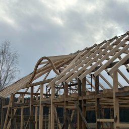 Michał Olechnowicz - Konstrukcja drewniana dachu z elementami łukowymi w trakcie budowy, widok na niebo z chmurami, częściowo pokryta łatami, drzewa w tle.