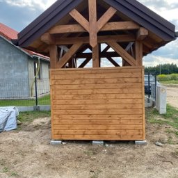 Michał Olechnowicz - Surowa drewniana konstrukcja altany z brązowym dachem, widoczna więźba dachowa, częściowo obudowana deskami, osadzona na betonowych bloczkach, na tle trawnika i domu w budowie.