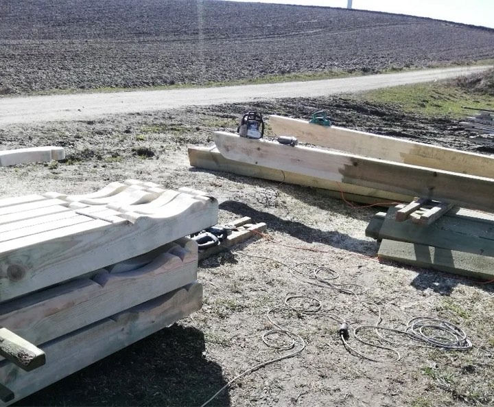 Przygotowane drewniane elementy konstrukcyjne z wyprofilowanymi zakończeniami, ułożone na ziemi obok piły łańcuchowej i przedłużaczy elektrycznych, w tle pole i droga polna.