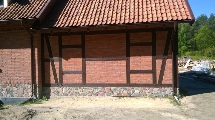 Fragment ściany budynku z czerwonej cegły z elementami konstrukcji szkieletowej z ciemnego drewna, widoczny dach z dachówki ceramicznej i rynna, w tle drzewa.