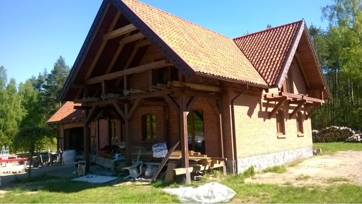 Dom w trakcie budowy: murowane ściany z cegły, drewniana konstrukcja dachu z widocznymi elementami więźby, czerwona dachówka, materiały budowlane na zewnątrz budynku, las w tle.