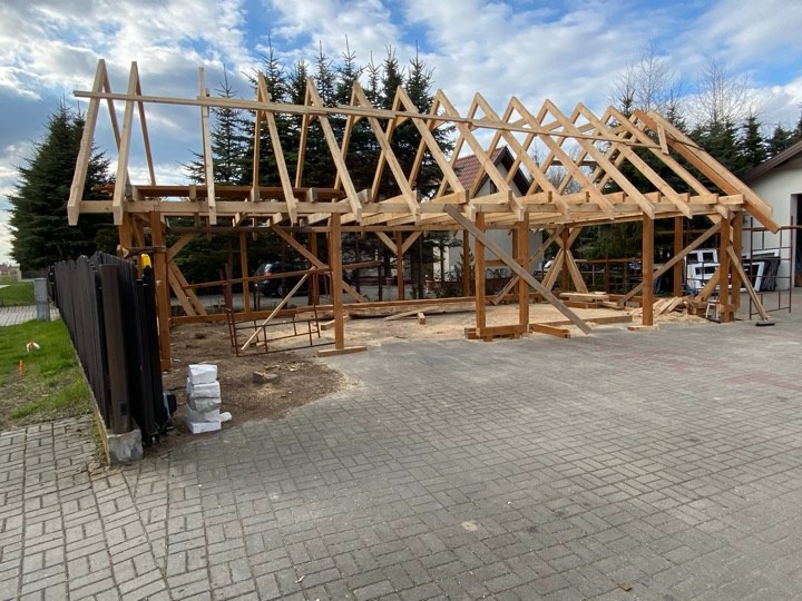 Wznoszenie drewnianej konstrukcji dachu na posesji, widoczna częściowo ukończona więźba dachowa z jasnego drewna, na pierwszym planie kostka brukowa, w tle drzewa iglaste i budynek.