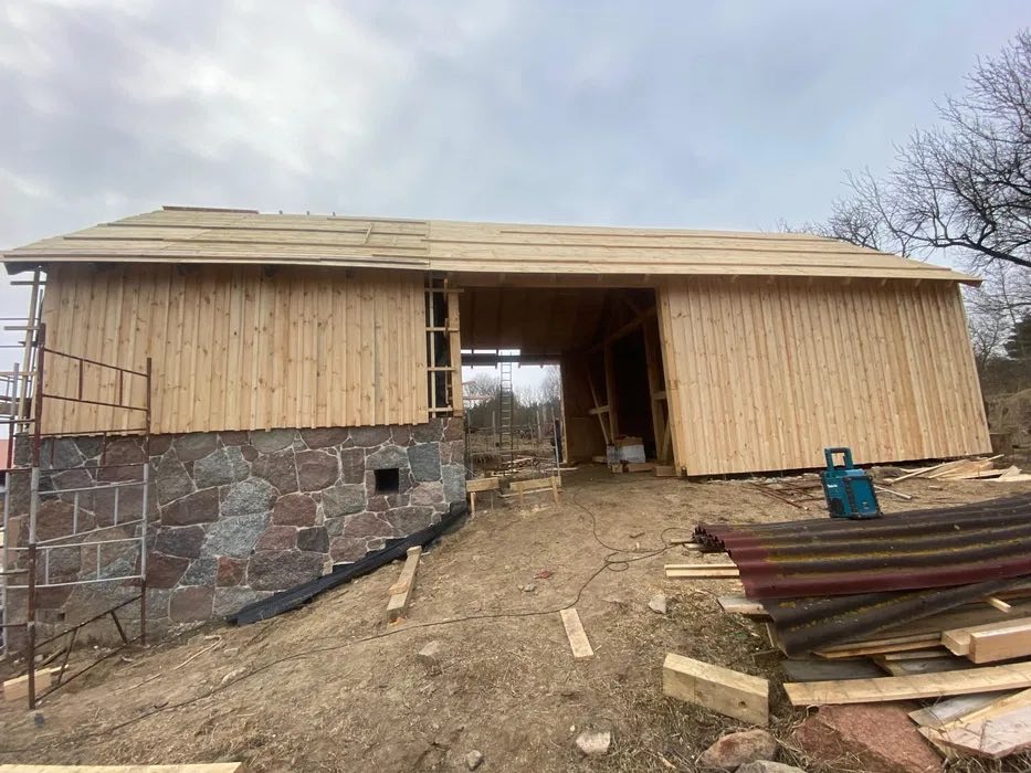 Budowa drewnianej stodoły z kamienną podmurówką w trakcie realizacji, widoczne rusztowania, deski na dachu i narzędzia.