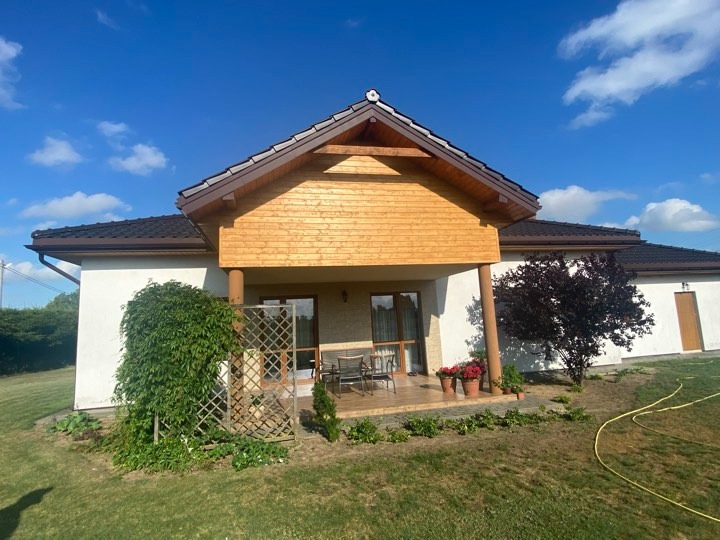 Dom jednorodzinny z ciemnobrązowym dachem i drewnianą elewacją werandy, widok z ogrodu w słoneczny dzień. W tle błękitne niebo z lekkimi chmurami. Na pierwszym planie trawnik i elementy roślinności.