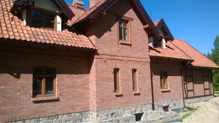 Dwupiętrowy dom z czerwonej cegły z terakotowym dachem, drewnianymi oknami i kamienną podmurówką, widziany z boku w słoneczny dzień.