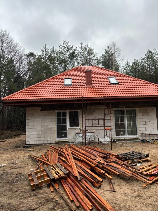 Dom w trakcie budowy z czerwonym dachem z dachówki ceramicznej, widoczne okna dachowe, komin i rusztowanie, na pierwszym planie stos drewna i palet.