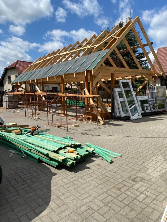 Wznoszenie drewnianej konstrukcji dachu z widoczną więźbą, pokrytą częściowo membraną dachową, na tle domów jednorodzinnych i zieleni, z przygotowanymi elementami drewnianymi i oknami PCV...