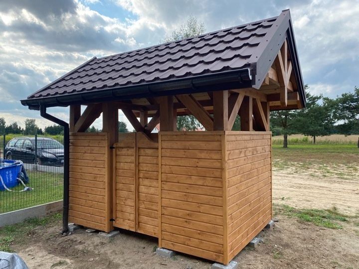 Drewniana altana ogrodowa z dwuspadowym dachem z brązowej blachodachówki, widoczny system rynnowy, na pierwszym planie ogrodzenie posesji, w tle samochód osobowy.