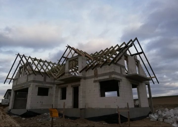 Surowy stan domu jednorodzinnego z widoczną konstrukcją drewnianą dachu, w tle pochmurne niebo.