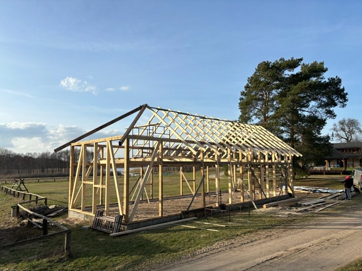 Konstrukcja drewniana dachu w trakcie budowy, widoczne elementy więźby dachowej na tle krajobrazu z drzewami i błękitnym niebem.