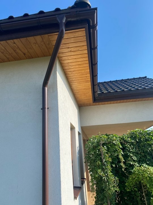 Dom jednorodzinny z brązowymi rynnami, drewnianą podbitką i ciemną dachówką w słoneczny dzień. Widoczny fragment elewacji z oknem i zielenią.