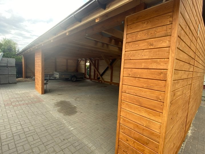 Drewniana wiata garażowa z widoczną konstrukcją więźby dachowej, pomalowana na brązowo, z przyczepką w środku, ujęcie z zewnątrz na tle kostki brukowej i stosu płyt.