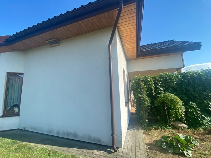 Fragment elewacji domu jednorodzinnego z brązowymi rynnami, drewnianą podbitką i ciemnym dachem. W oknie widoczny kot.