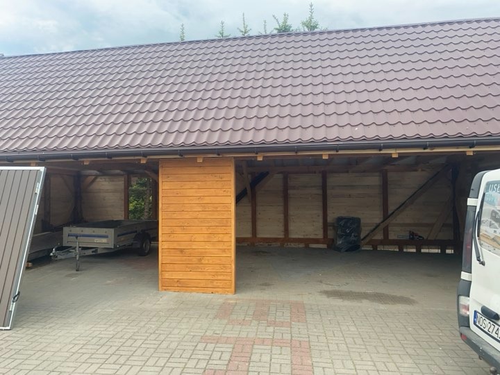 Zadaszenie z brązową blachodachówką, drewniana konstrukcja, przesuwne panele z desek, przyczepka samochodowa wewnątrz, widok z poziomu kostki brukowej.