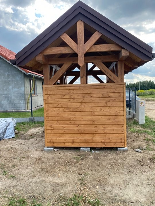 Surowa drewniana konstrukcja altany z brązowym dachem, widoczna więźba dachowa, częściowo obudowana deskami, osadzona na betonowych bloczkach, na tle trawnika i domu w budowie.