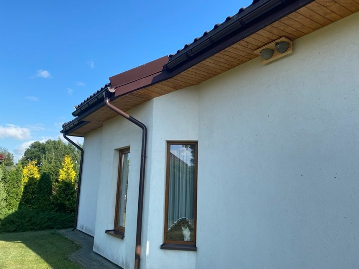 Dom jednorodzinny z brązowymi rynnami i obróbkami blacharskimi, widok z zewnątrz, w oknie widoczny kot.