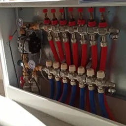 Rozdzielacz podłogowy z pompą obiegową i manometrami, z widocznymi rurami w kolorach czerwonym i niebieskim, zamontowany w szafce wnękowej.