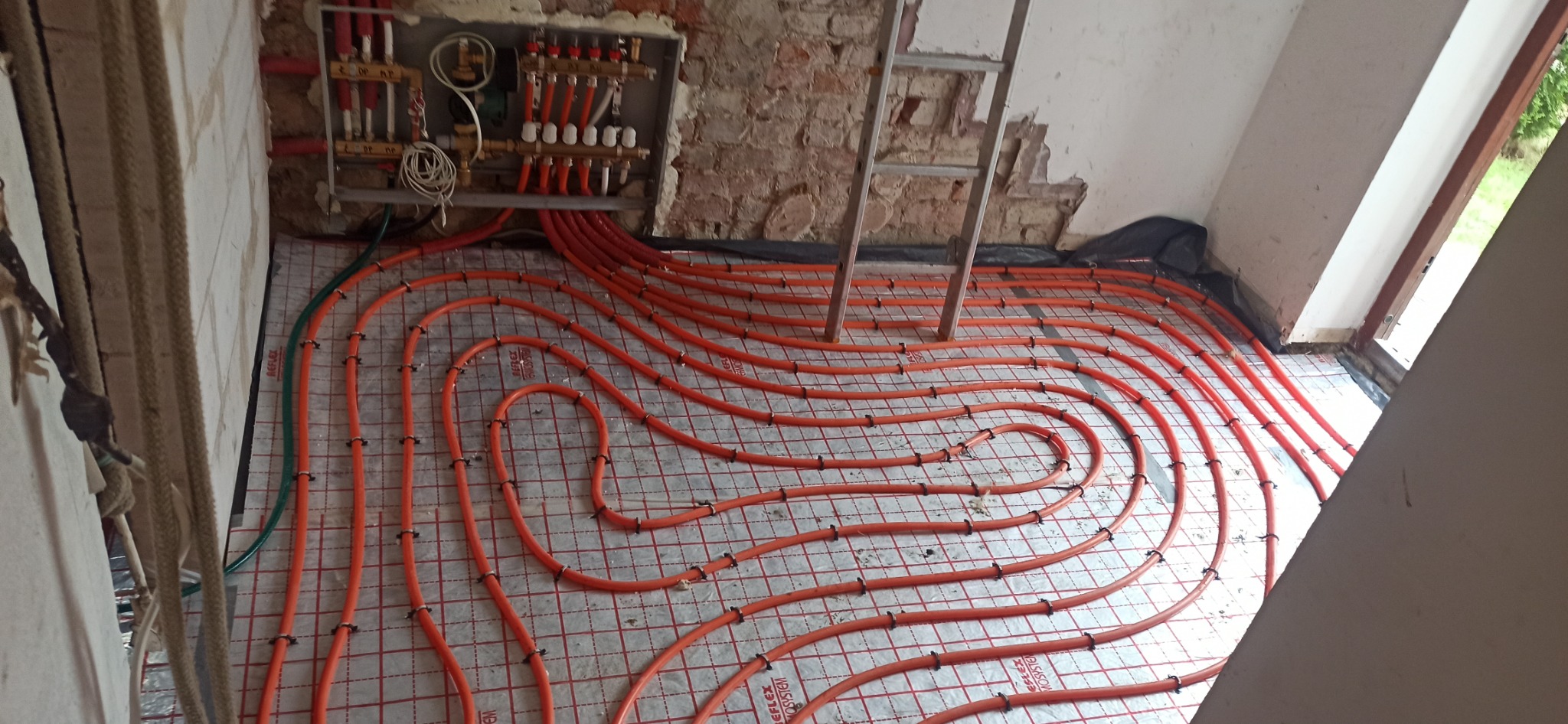 Montaż ogrzewania podłogowego: pomarańczowe rury grzewcze ułożone na izolacji z siatką, widoczny rozdzielacz z zaworami i drabina oparta o ścianę z fragmentami cegieł.
