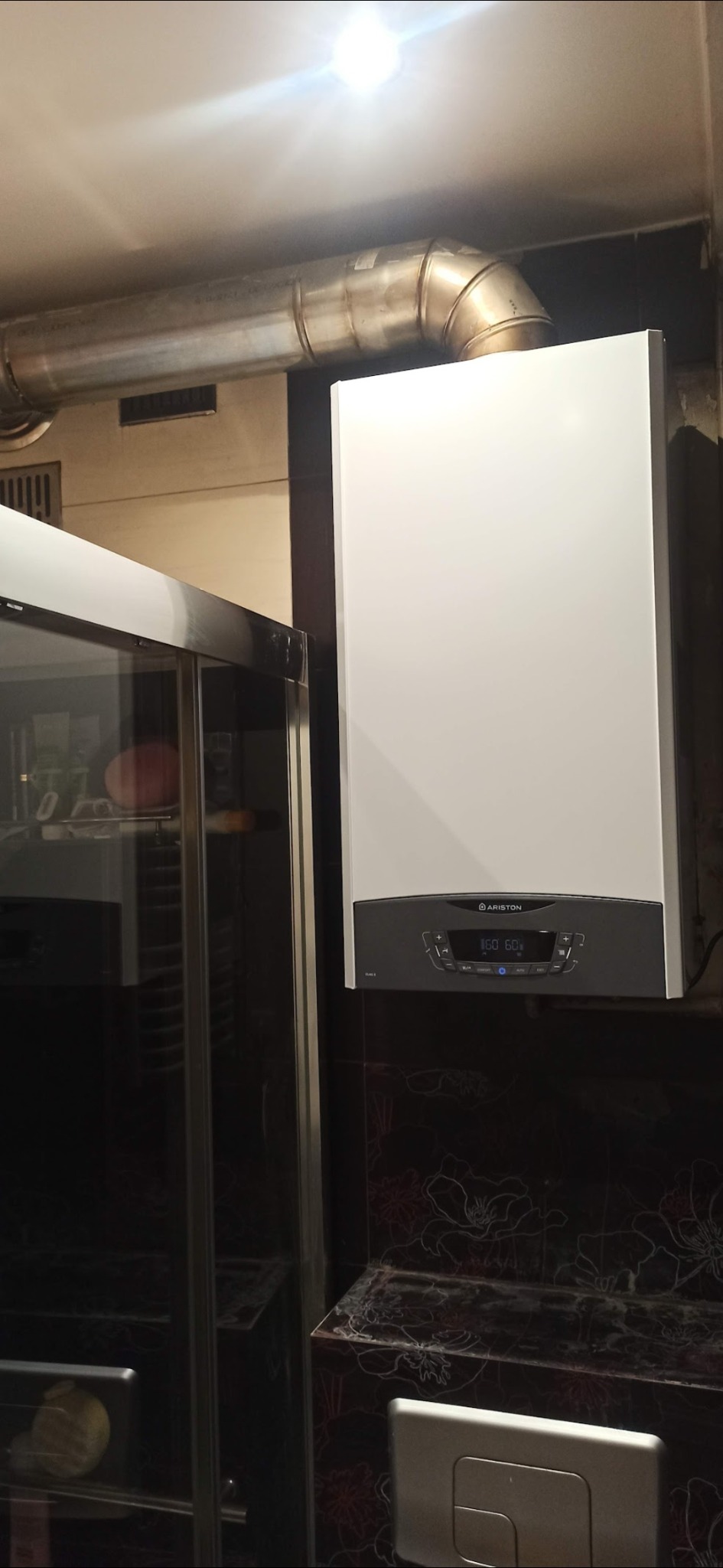 Biały piec gazowy Ariston zamontowany na ścianie łazienki z ciemnymi, kwiatowymi płytkami, widoczny fragment rury odprowadzającej spaliny i kabina prysznicowa.