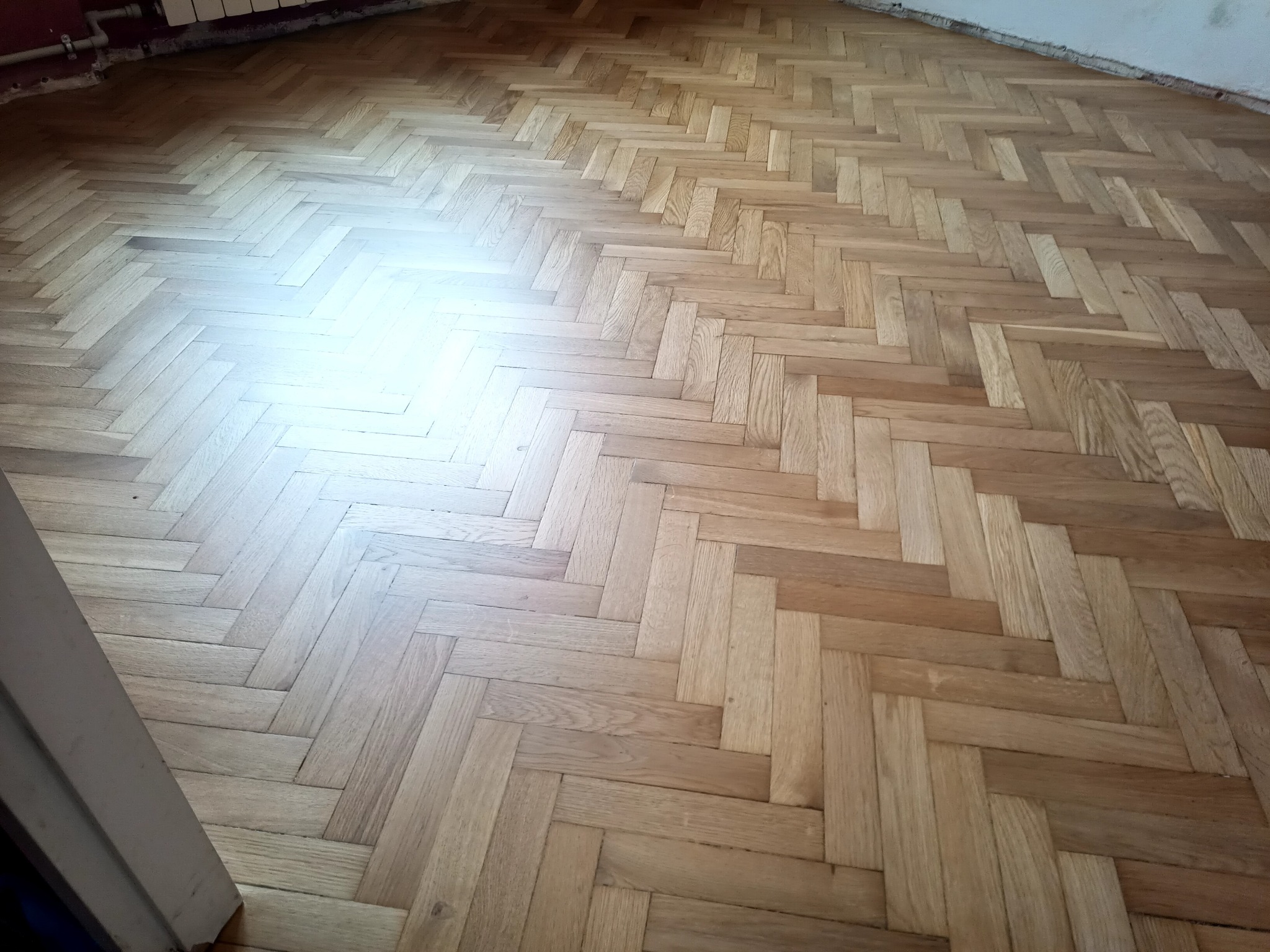 Parkiet ułożony w jodełkę, po renowacji, z widocznym połyskiem w centralnej części, blisko ściany widoczne niedoskonałości.