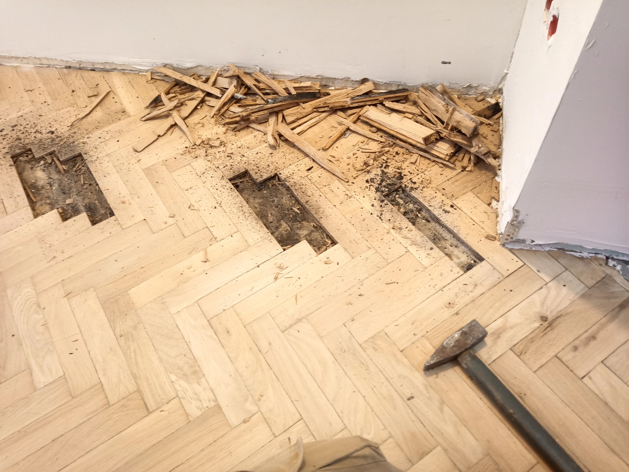 Fragment podłogi parkietowej w trakcie naprawy, z widocznymi ubytkami, stertą odłamków drewna przy ścianie oraz leżącym obok młotkiem