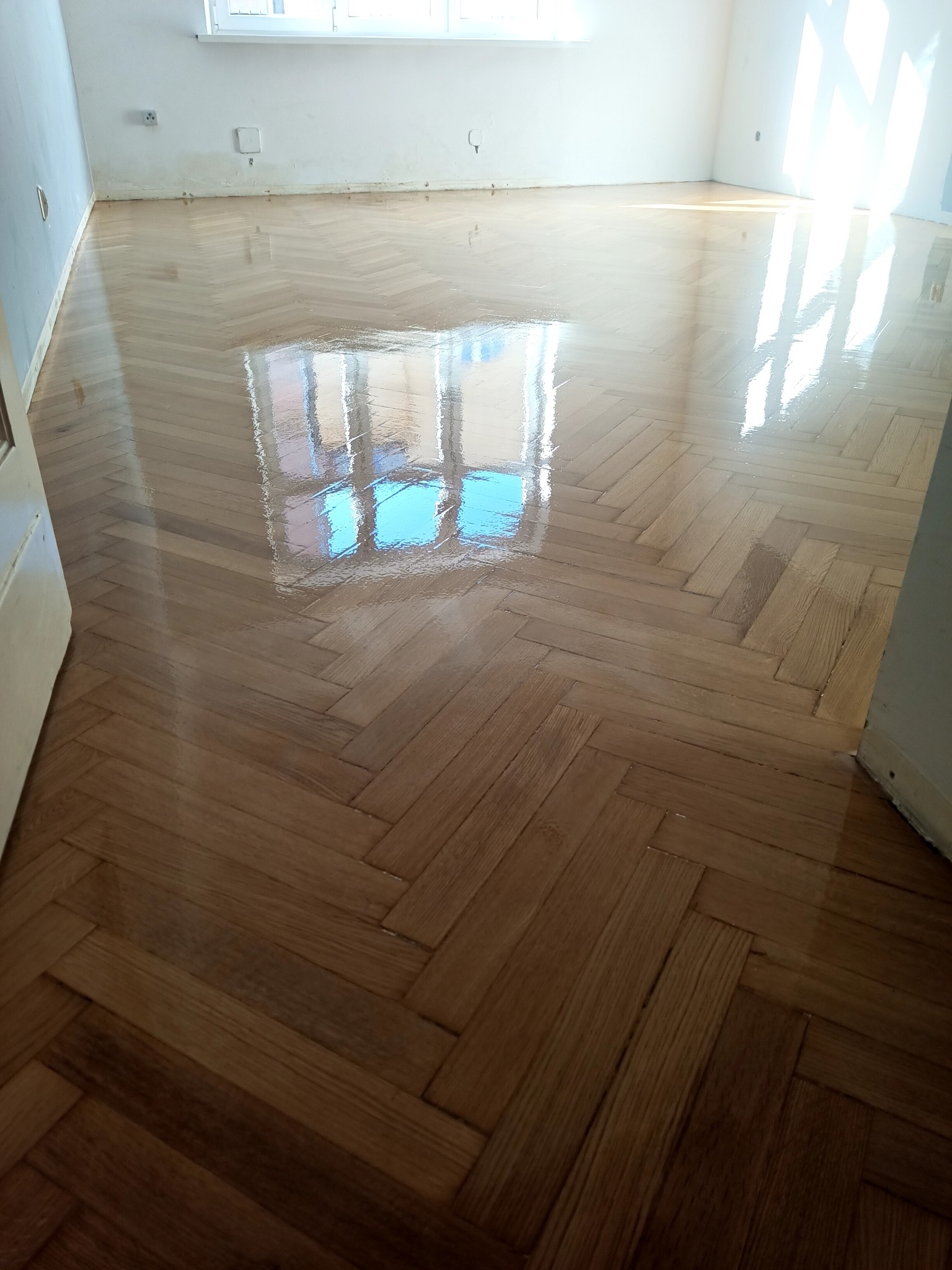 Świeżo polakierowany parkiet ułożony w jodełkę, odbijający światło okna, widoczne drobne niedoskonałości na ścianie.