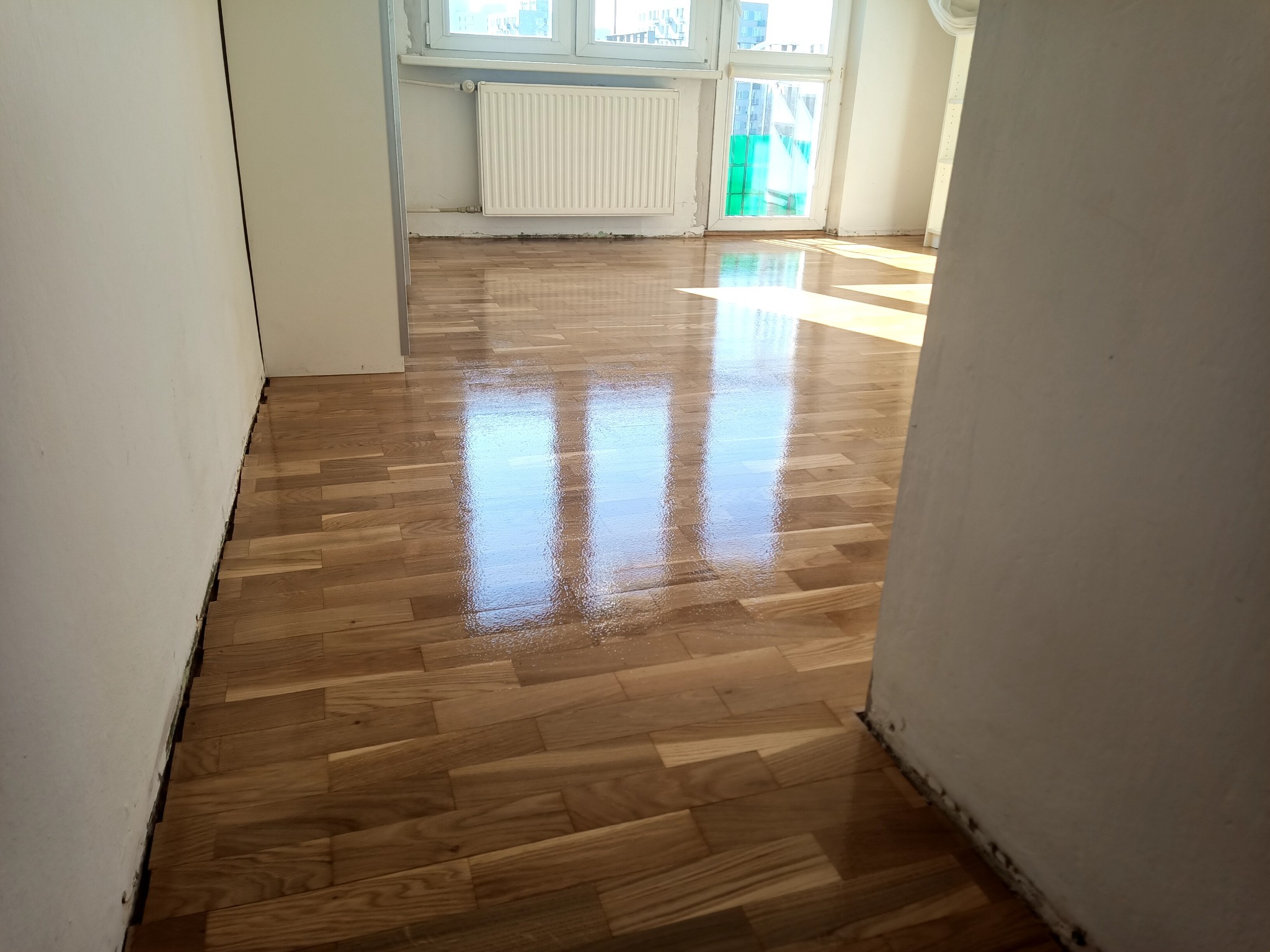 Błyszcząca, świeżo polakierowana podłoga parkietowa z widocznymi refleksami światła słonecznego w pustym pokoju z białymi ścianami i grzejnikiem pod oknem.