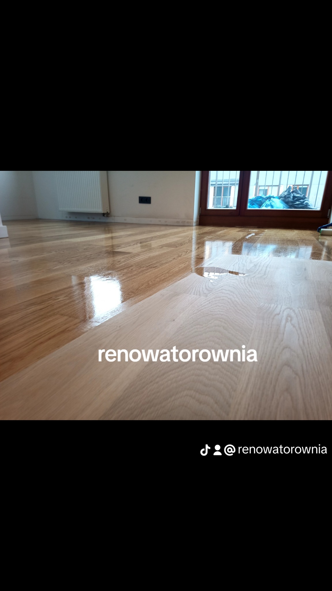 Częściowo wycyklinowana, błyszcząca drewniana podłoga w pokoju z grzejnikiem i oknem balkonowym w tle.