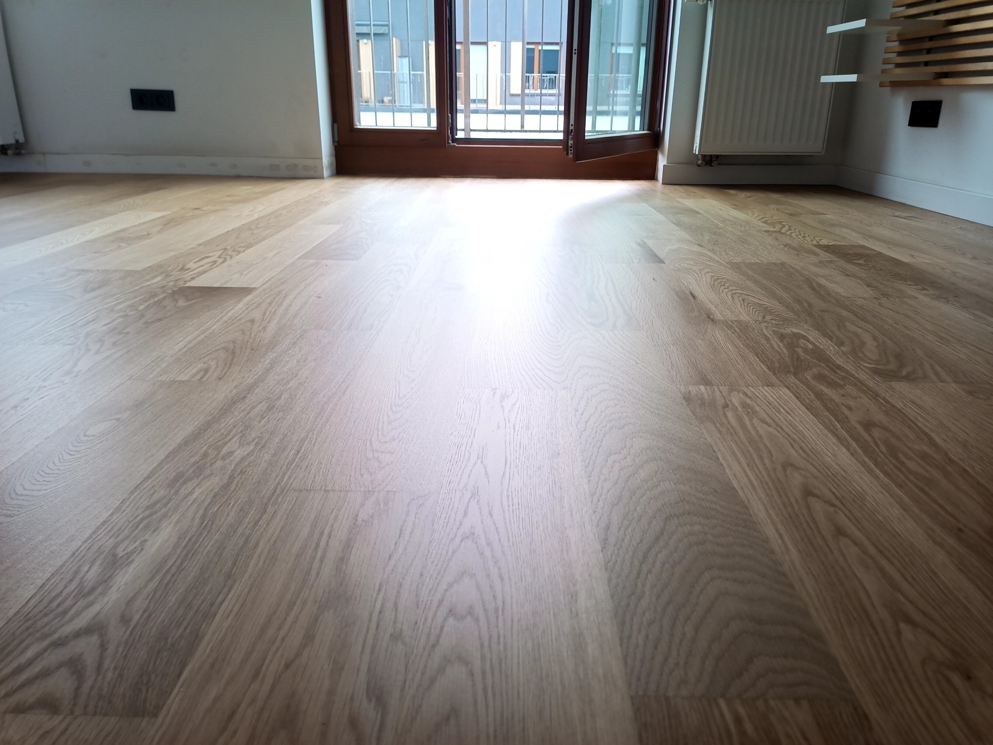 Jasna, odnowiona podłoga drewniana w pokoju z otwartym balkonem, widoczne detale usłojenia drewna i listwy przypodłogowe.