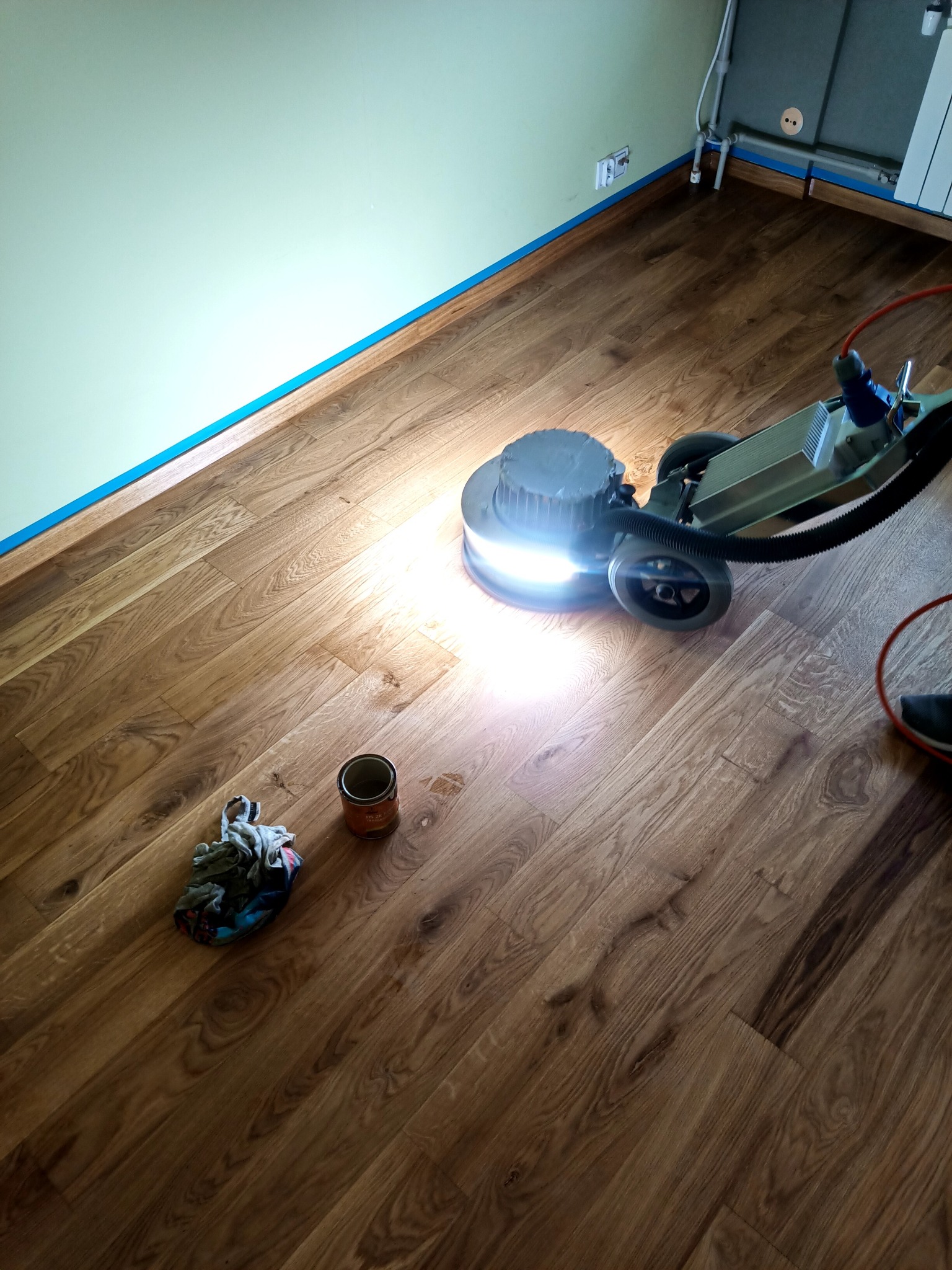Maszyna do polerowania parkietu w trakcie pracy, jasne światło z maszyny oświetla drewnianą podłogę, widoczna puszka lakieru i szmatka obok, ściany zabezpieczone niebieską taśmą.