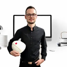 Mężczyzna w okularach i czarnej koszuli trzymający biały kask ochronny na tle biurka z lampką, monitorem i czajnikiem.