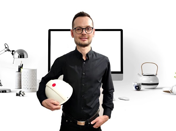 Mężczyzna w okularach i czarnej koszuli trzymający biały kask ochronny na tle biurka z lampką, monitorem i czajnikiem.