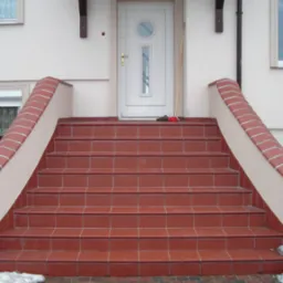 Zewnętrzne schody wejściowe do domu wyłożone czerwoną terakotą z jasnymi fugami, zakończone obrzeżem z cegieł klinkierowych, widoczne ślady śniegu.