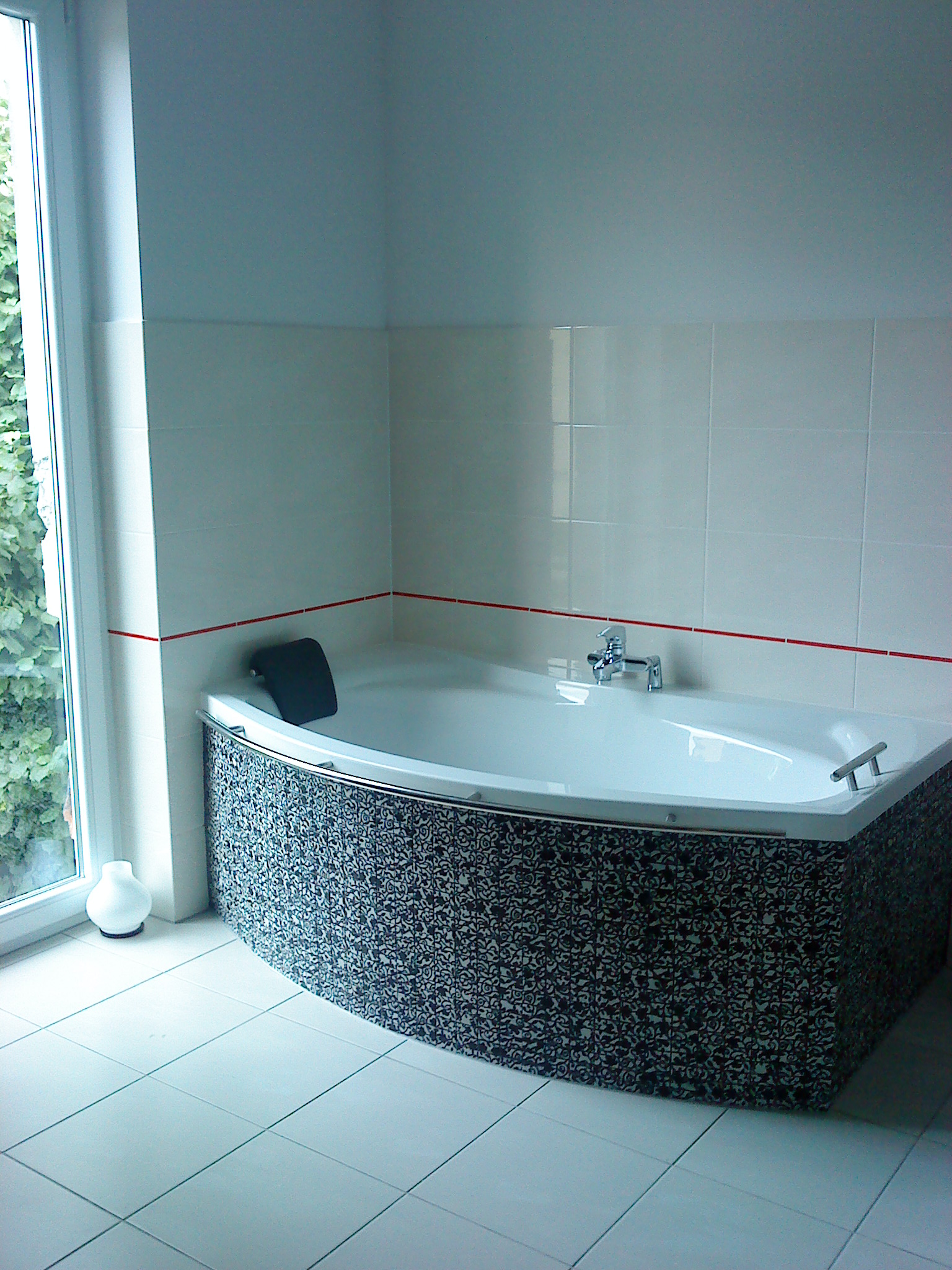 Narożna wanna z hydromasażem, obudowana mozaiką w czarno-białe wzory, z chromowaną armaturą i zagłówkiem, na tle białych płytek z czerwoną listwą, obok okna z widokiem na zieleń.