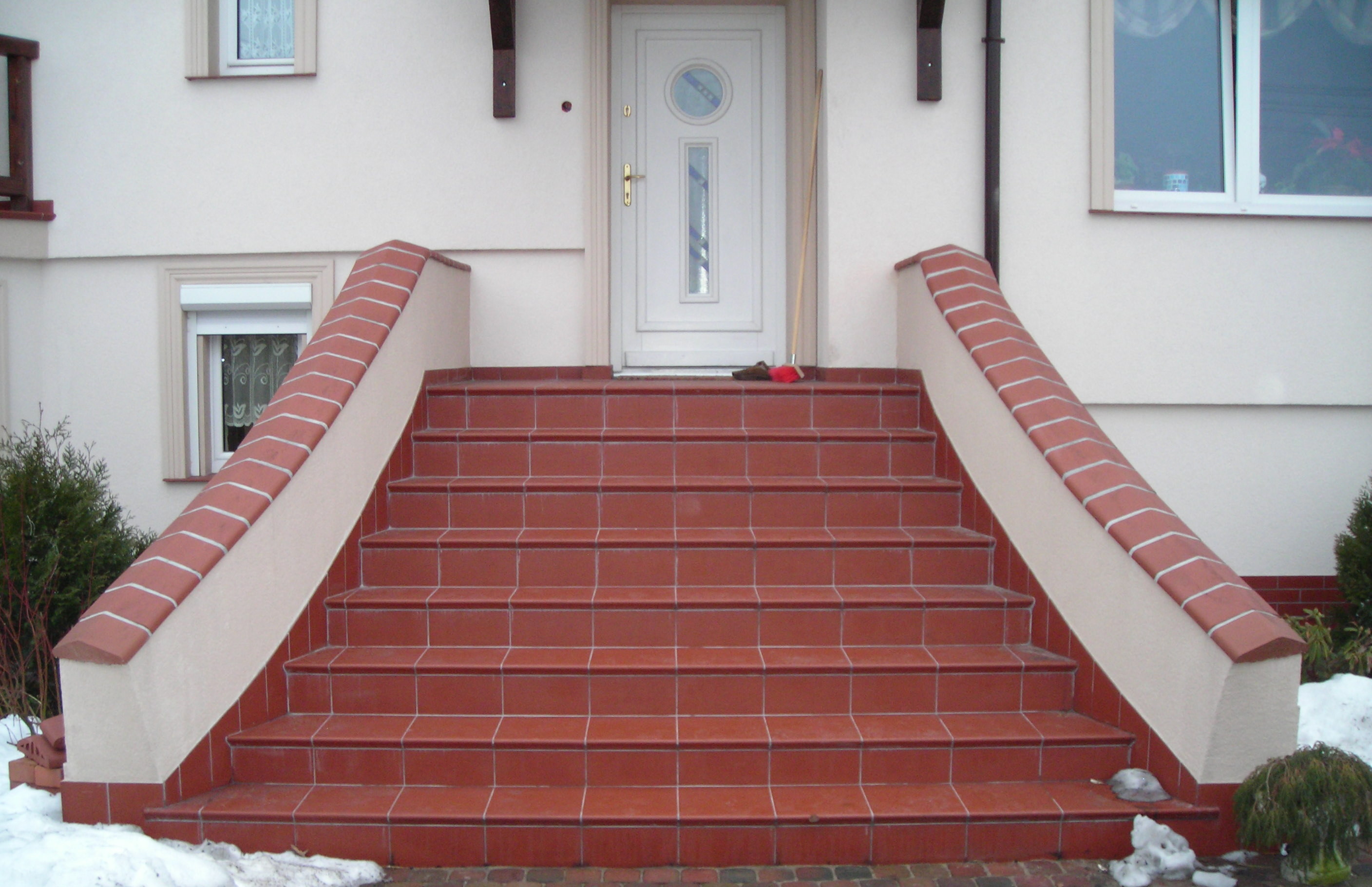 Zewnętrzne schody wejściowe do domu wyłożone czerwoną terakotą z jasnymi fugami, zakończone obrzeżem z cegieł klinkierowych, widoczne ślady śniegu.