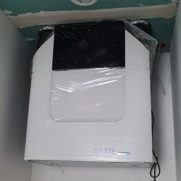 Serhii Trokhin - Biała jednostka rekuperacyjna marki Aeris, jeszcze w folii ochronnej, zamontowana pod sufitem w pomieszczeniu w trakcie wykańczania, widoczne przewody elektryczne i niedokończone połączenia.