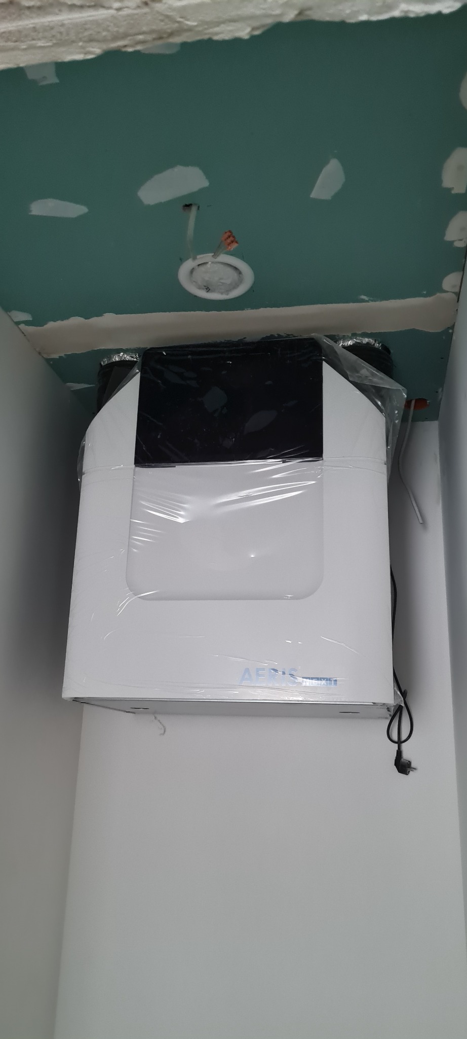 Biała jednostka rekuperacyjna marki Aeris, jeszcze w folii ochronnej, zamontowana pod sufitem w pomieszczeniu w trakcie wykańczania, widoczne przewody elektryczne i niedokończone połączenia.