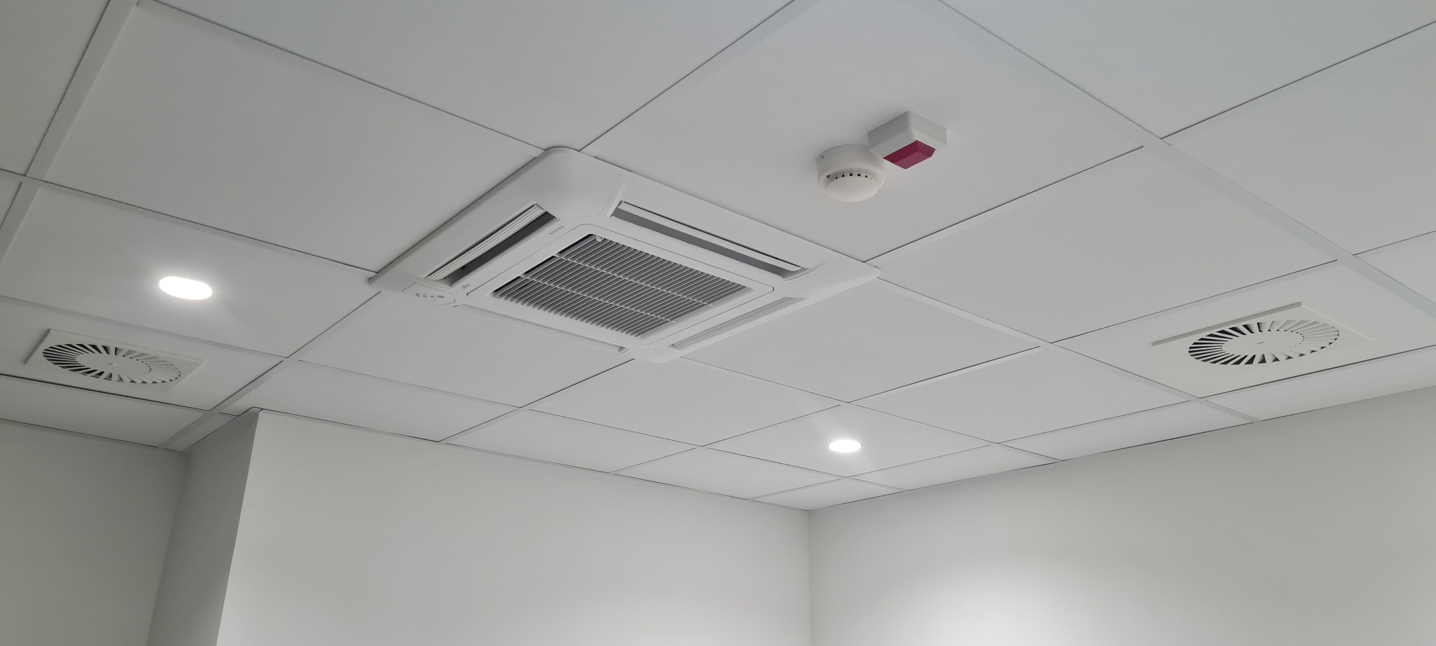Kasetonowa klimatyzacja sufitowa w biurze, widoczne kratki wentylacyjne i czujnik dymu na białym suficie podwieszanym.