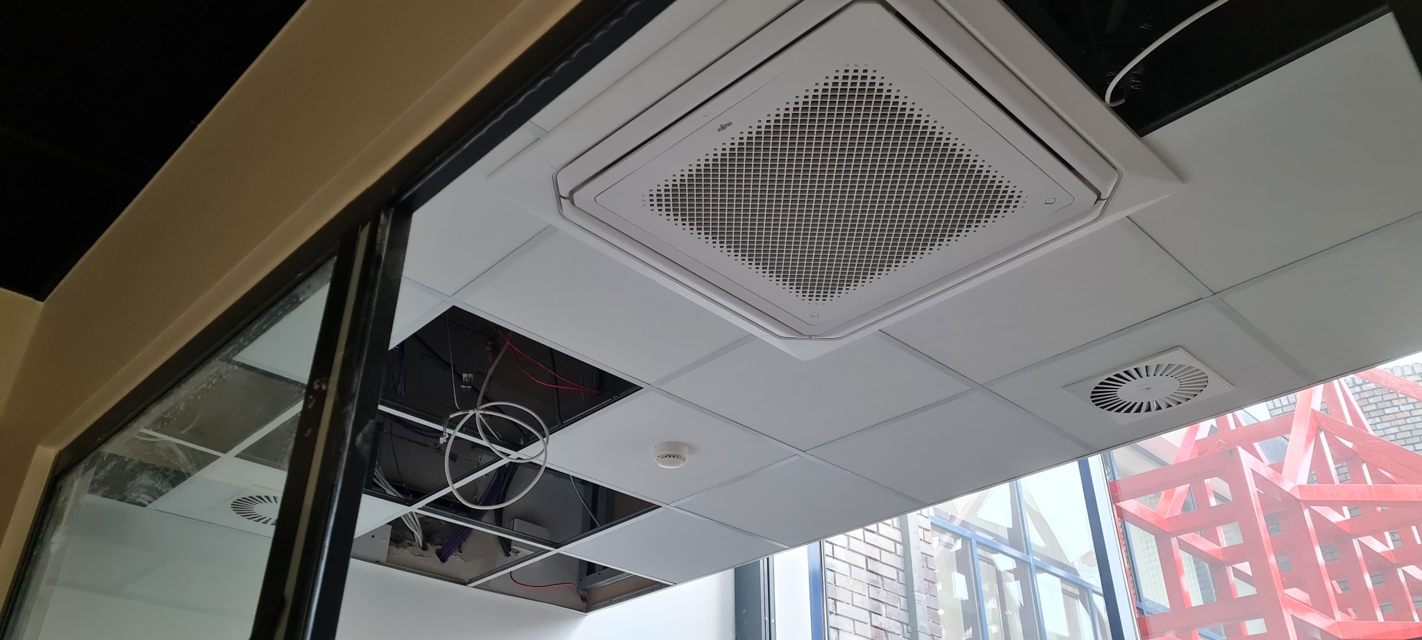 Sufit kasetonowy z zamontowaną jednostką klimatyzacji Fujitsu, widoczne otwory w suficie z instalacją elektryczną i wentylacyjną, w tle konstrukcja rusztowania za oknem.