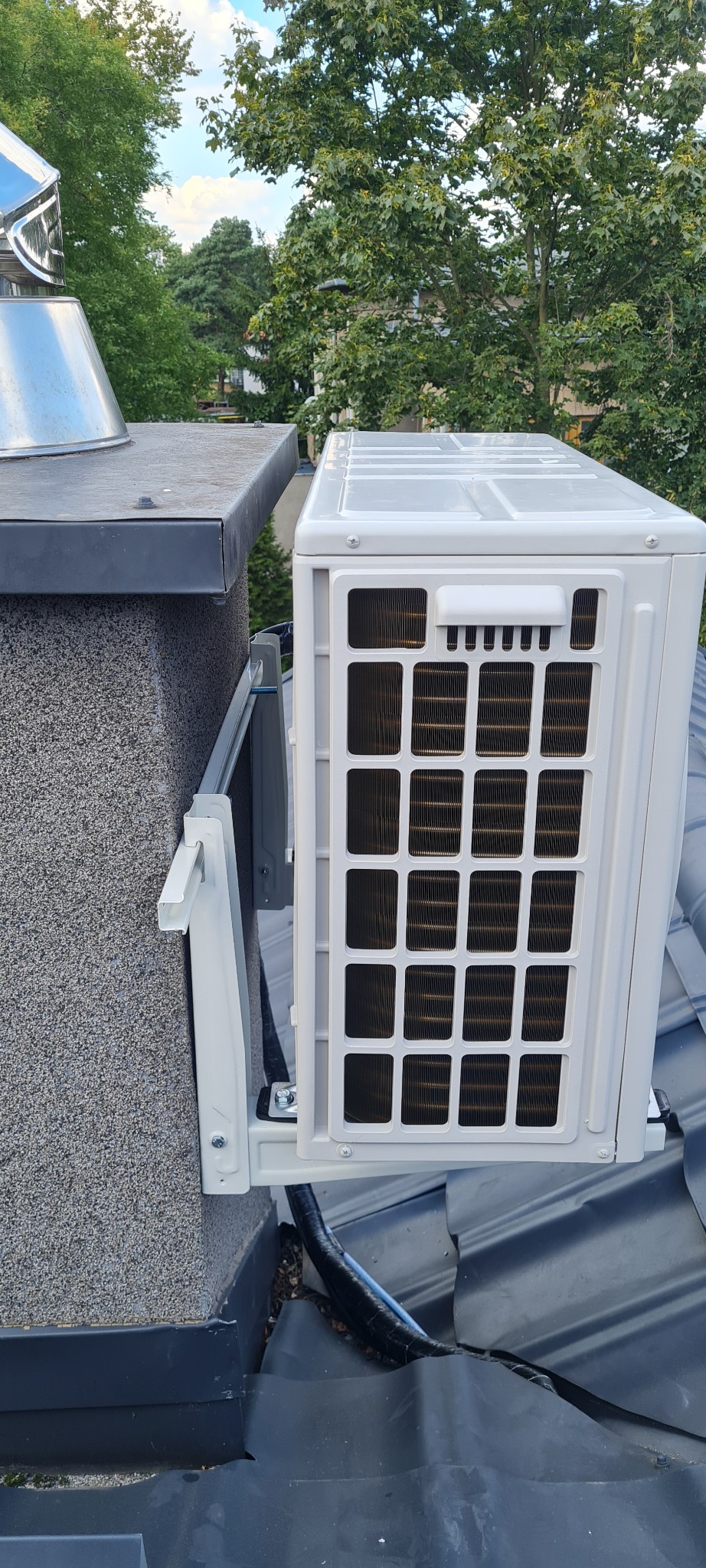 Zewnętrzna jednostka klimatyzacji zamontowana na dachu budynku, widok z bliska na szarej dachówce i obok komina z metalowym zakończeniem, na tle zieleni drzew.