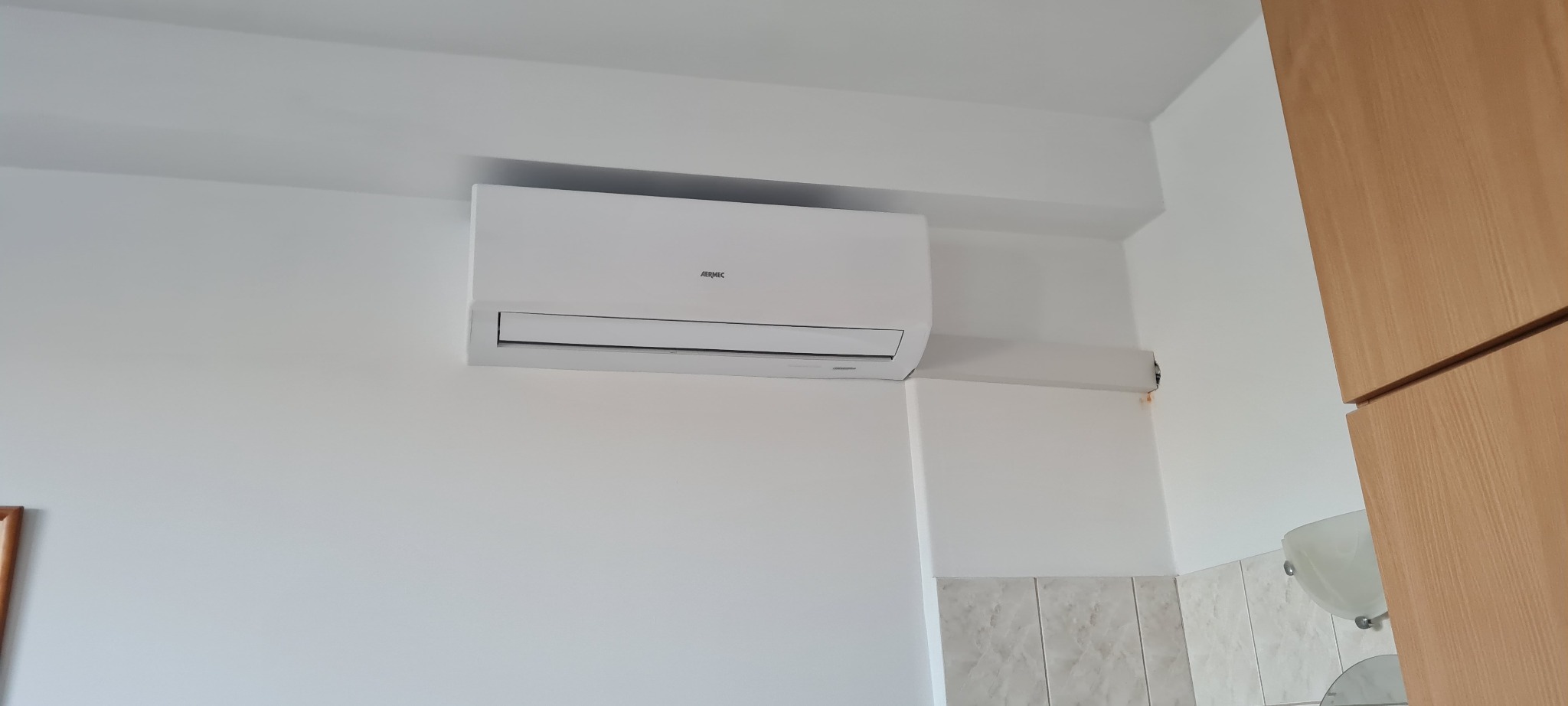 Biały klimatyzator Aermec zamontowany na ścianie w pomieszczeniu z jasnymi płytkami i drewnianymi szafkami.