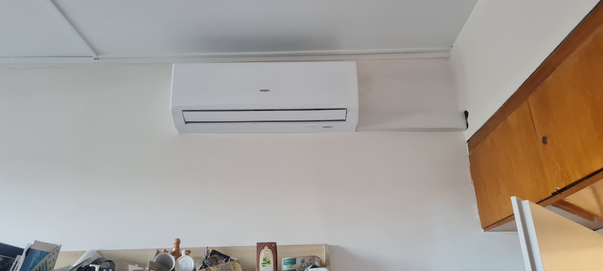 Biała jednostka klimatyzacji Aermec zamontowana na ścianie w pomieszczeniu z widoczną szafką kuchenną w rogu.