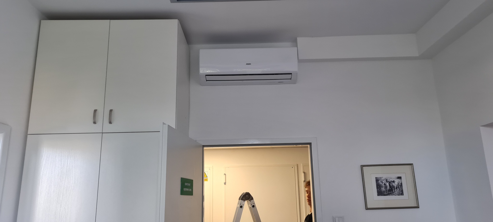 Biały klimatyzator ścienny zamontowany nad drzwiami w jasnym pomieszczeniu biurowym, widoczna drabina i szafka w tle.