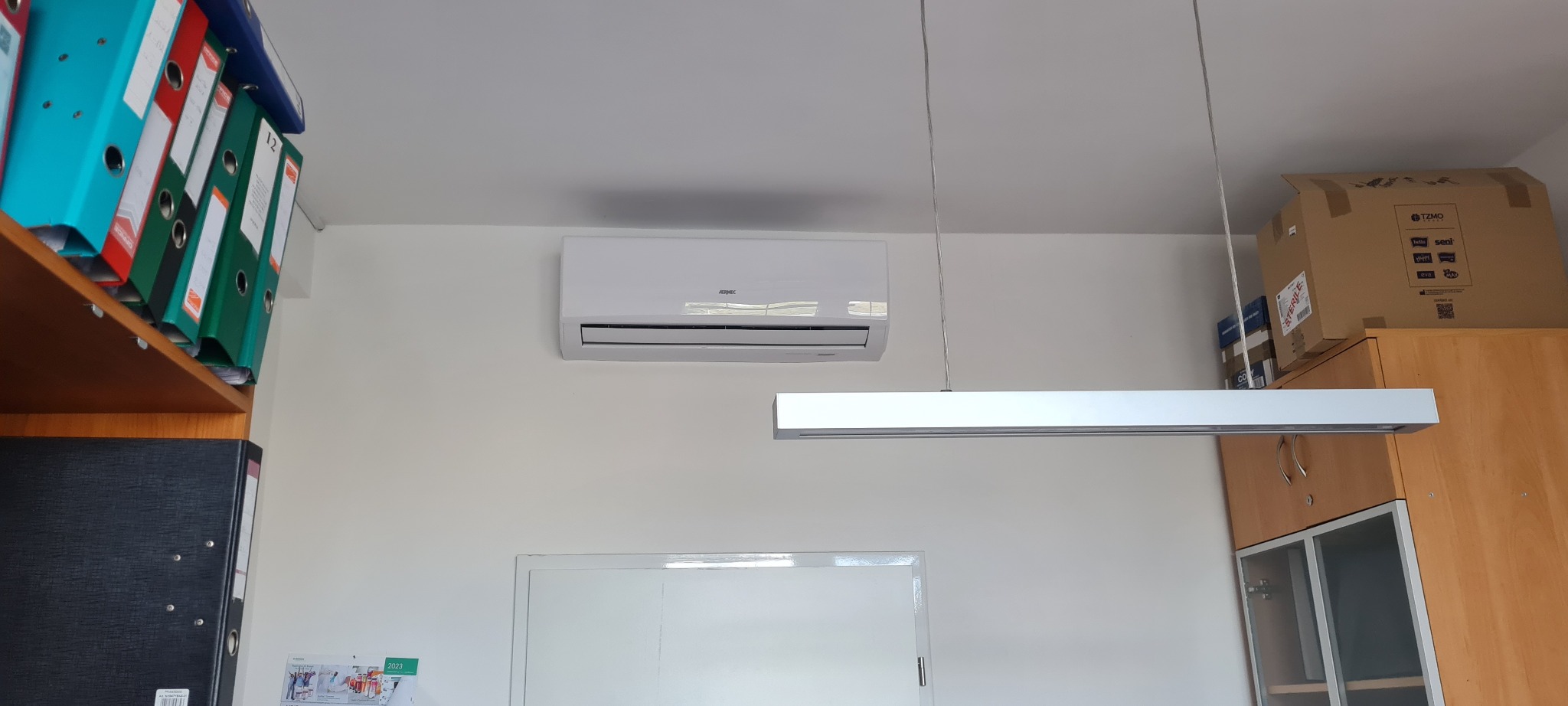 Biała jednostka klimatyzacyjna zamontowana na ścianie w biurze, widoczna również lampa sufitowa i regał z segregatorami
