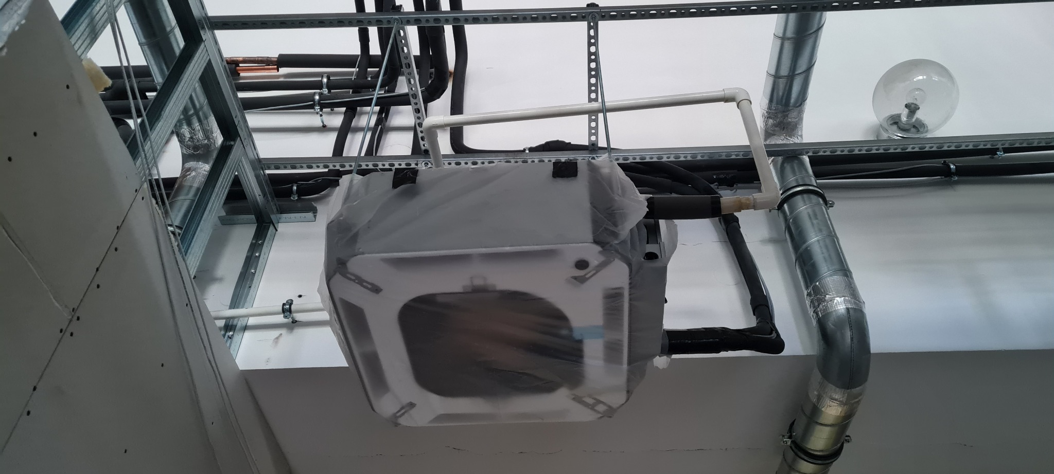 Klimatyzator kasetonowy w folii ochronnej, zawieszony pod sufitem podwieszanym w trakcie montażu, widoczne rury i instalacje.