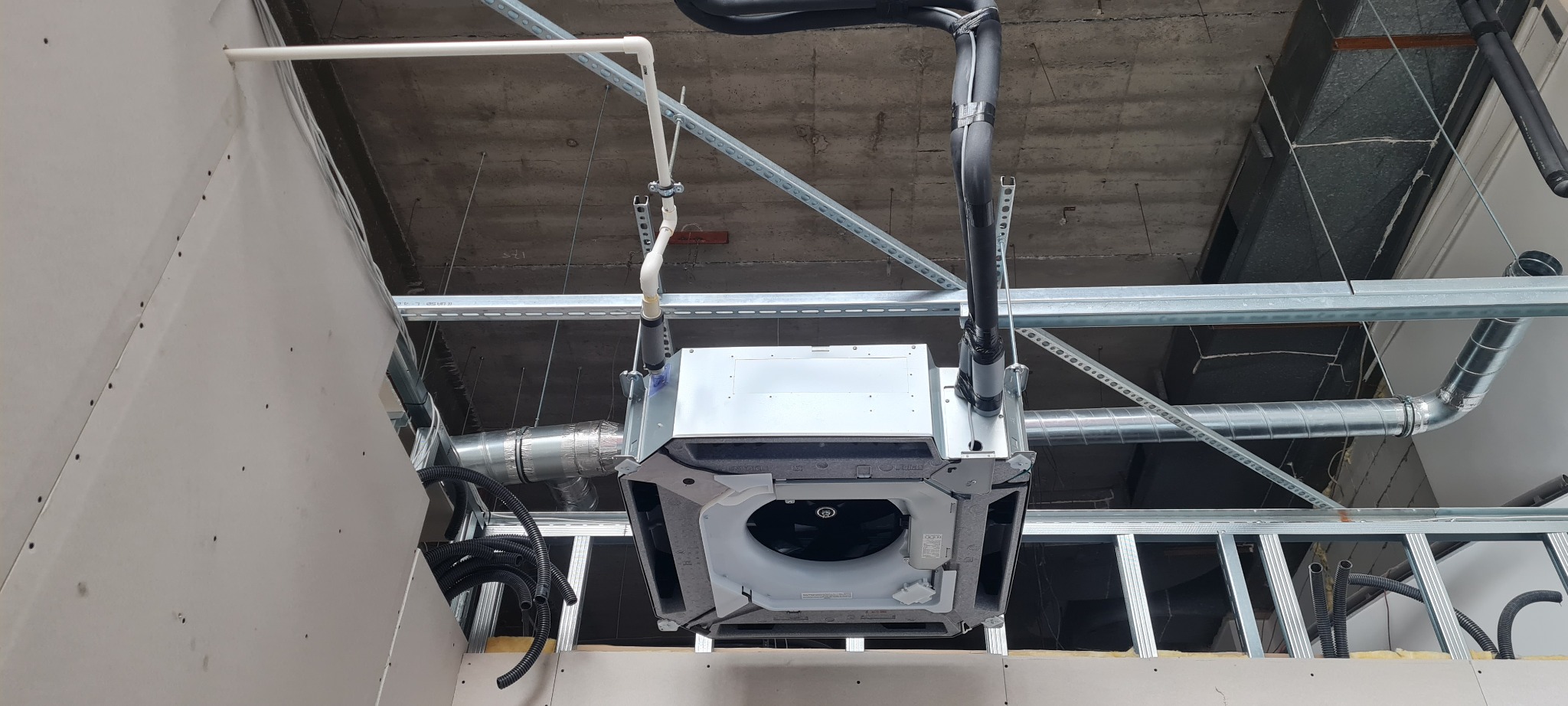 Montaż podwieszanej jednostki klimatyzacyjnej w surowym wnętrzu, widoczne rury i przewody wentylacyjne, konstrukcja sufitu podwieszanego w tle.