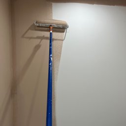 Kolorowy Kąt - Malowanie ściany wałkiem na teleskopowym kiju. Częściowo pomalowana ściana w odcieniach beżu z niebieską taśmą malarską przy suficie.